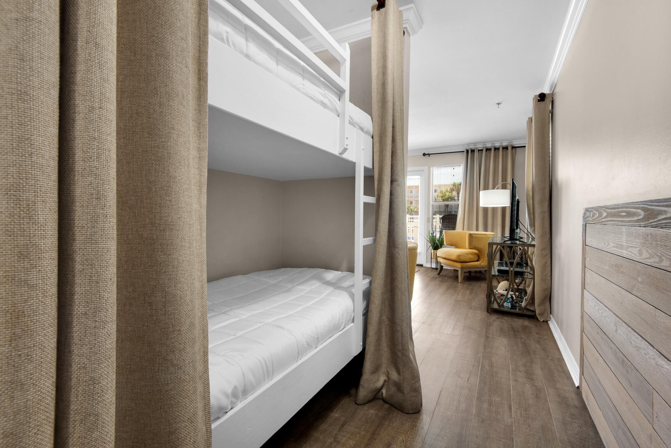 A hotel room with a bunk bed, a small table with a lamp, a chair, and a view of the outside.