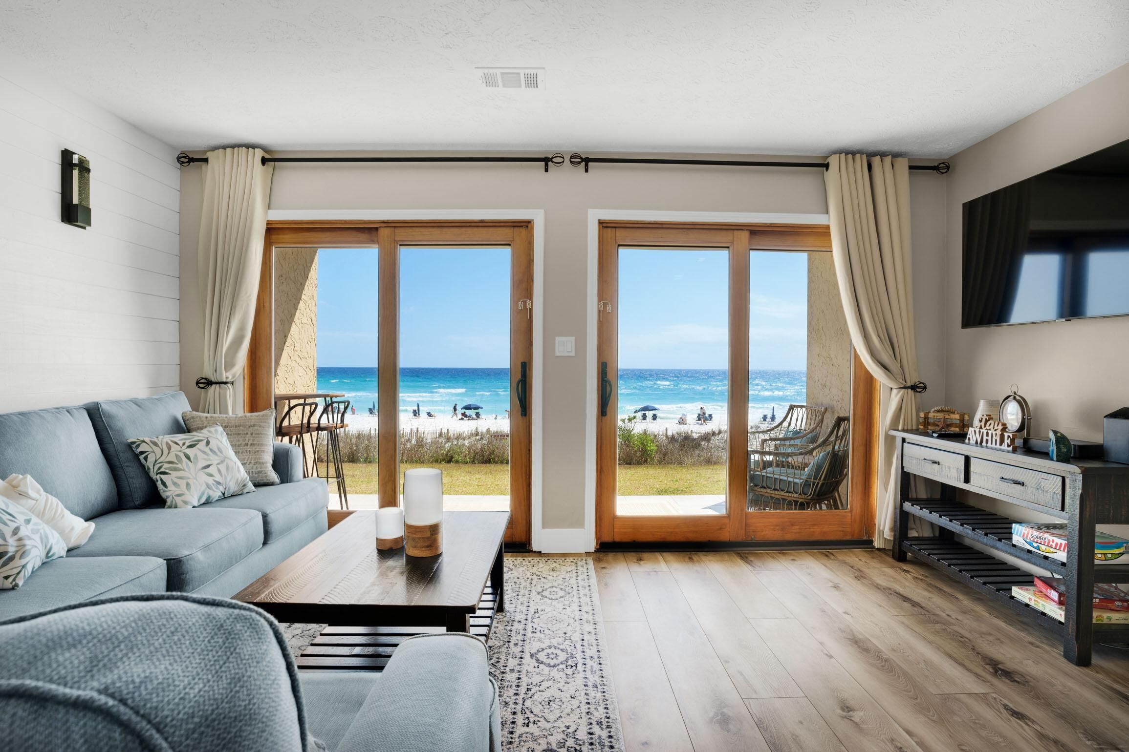 Ein modernes Wohnzimmer mit einem großen Schiebeglastür, die Blick auf einen Strand bietet, einer Couch, einem Couchtisch und einem an der Wand befestigten Fernseher.