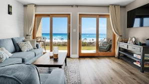 A modern living room with a large sliding glass door leading to a beach view, a sofa, a coffee table, and a TV on the wall. - Crystal Villas A-3 (Destin)