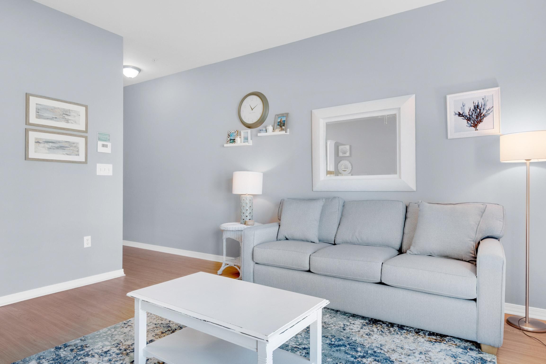 Un salon avec un canapé gris clair, une table basse blanche et un tapis à motifs bleus. On y trouve une horloge murale, des tableaux encadrés et un lampadaire.