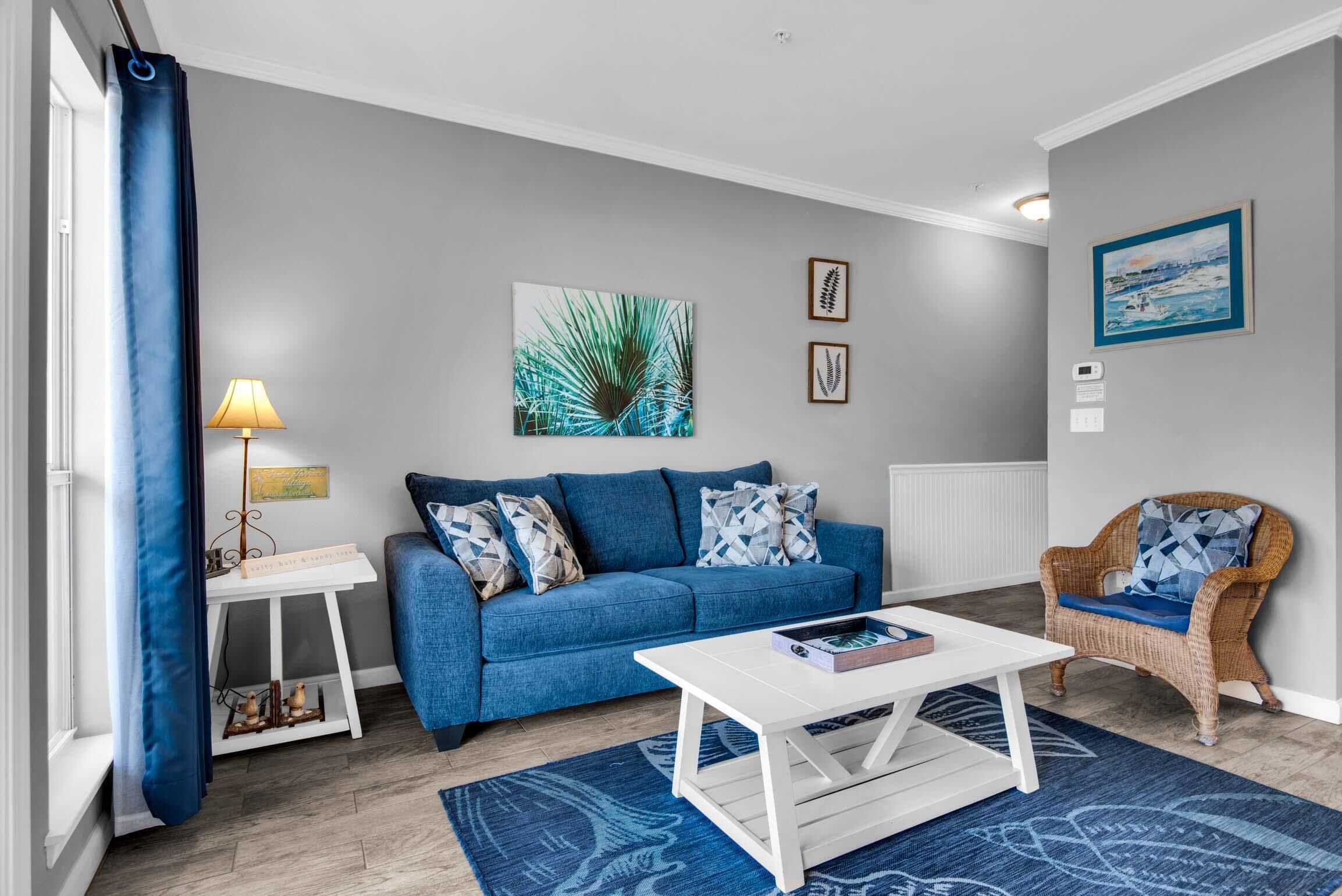Una sala de estar con un sofá azul, una mesa de centro blanca y una silla de mimbre. Hay un cuadro en la pared y una lámpara sobre una mesita auxiliar.