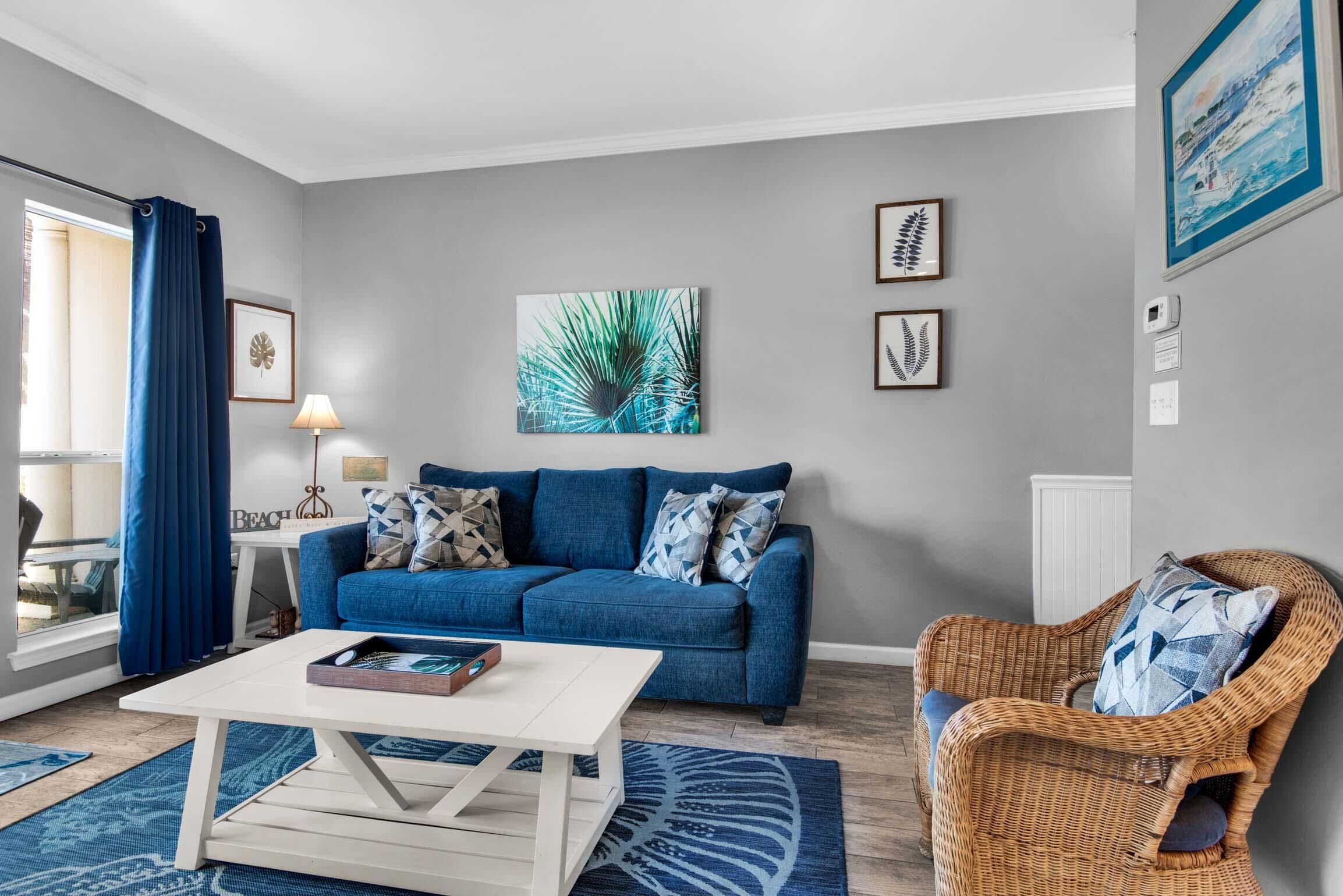 Ein Wohnzimmer mit einem blauen Sofa, einem Korbstuhl und einem weißen Couchtisch. An der Wand hängen zwei gerahmte Kunstwerke und ein großes Fenster mit Vorhängen.