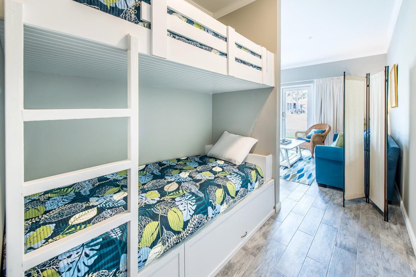 Una cama litera con colcha floral, una mesita de noche pequeña y una silla en una habitación con vistas al exterior.