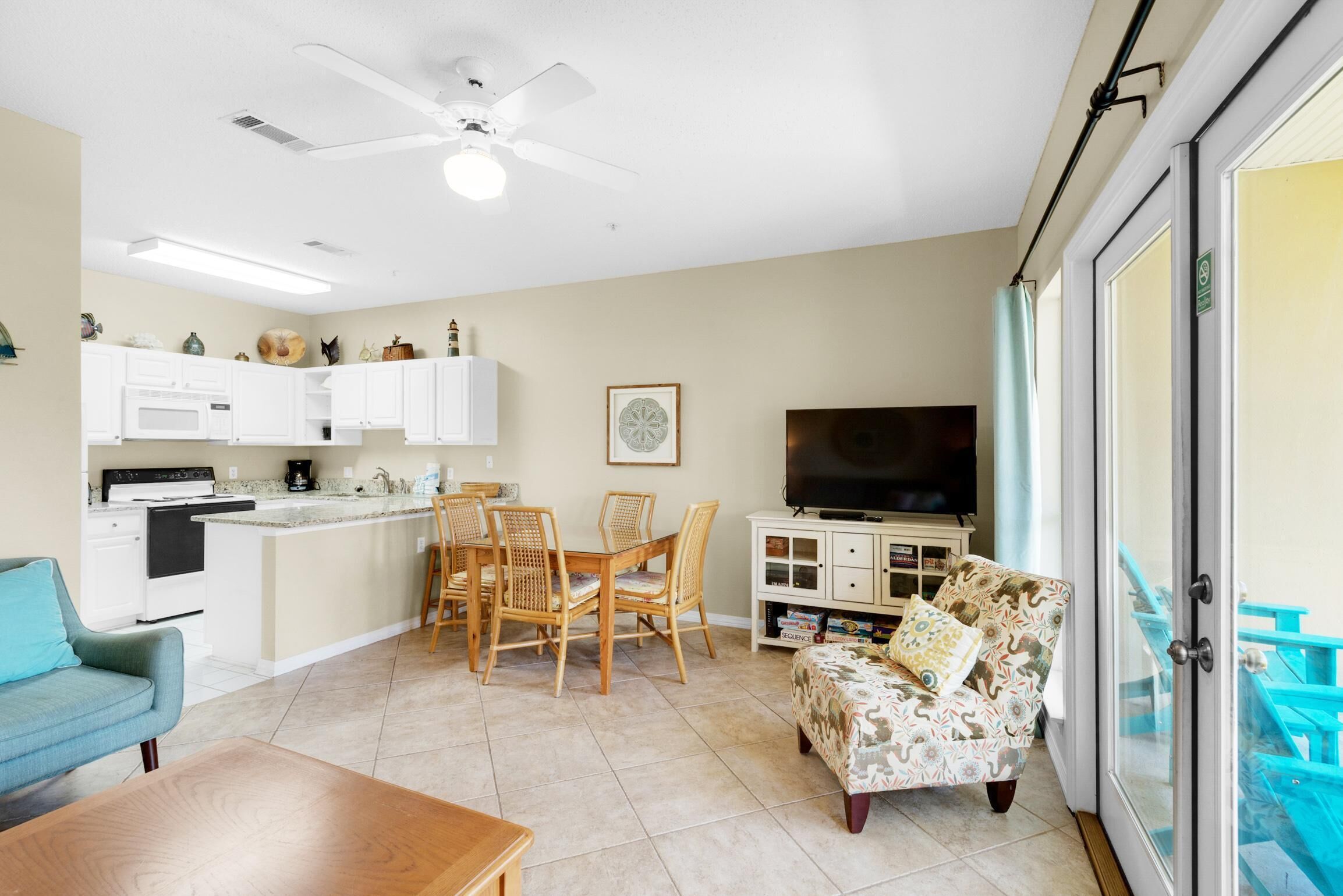 A modern living room with a sofa, coffee table, and a TV. There is a dining area with a table and chairs. The room has a ceiling fan and a large window with a view.