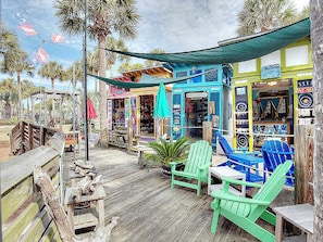 A wooden deck with green Adirondack chairs, a small table, and a tiki bar with colorful decorations. - Whispering Palms (Santa Rosa Beach)