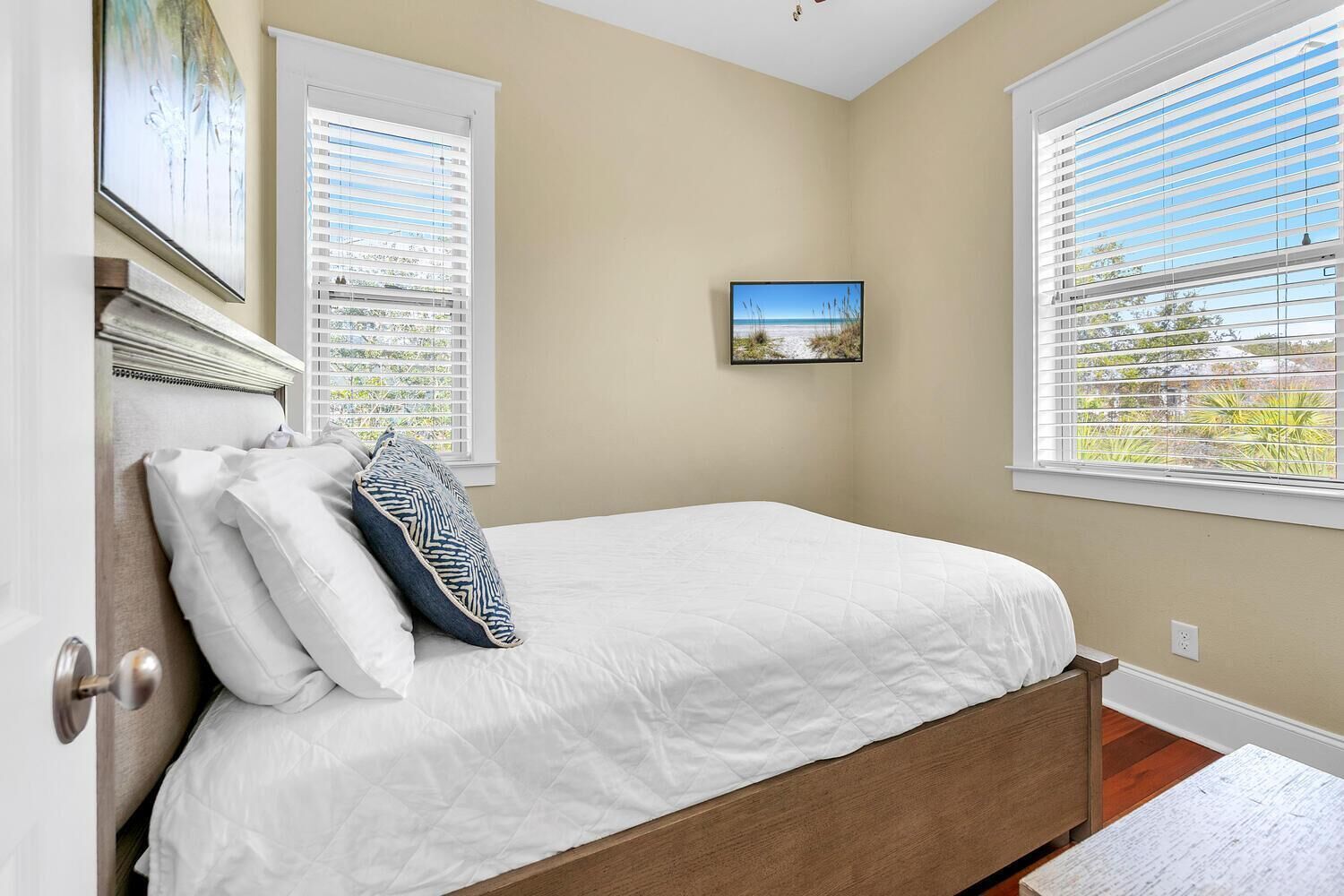 A bedroom with a bed, two windows with blinds, a painting on the wall, and a door.
