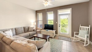 A living room with a beige sofa, a coffee table, a TV on a cabinet, a rocking chair, and a ceiling fan. - Topsail Village 422 (Santa Rosa Beach)