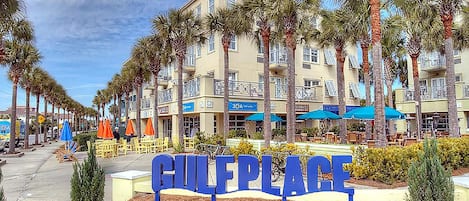 Letrero que dice 'GULFPLACE', con una fila de palmeras y una hilera de edificios al fondo.