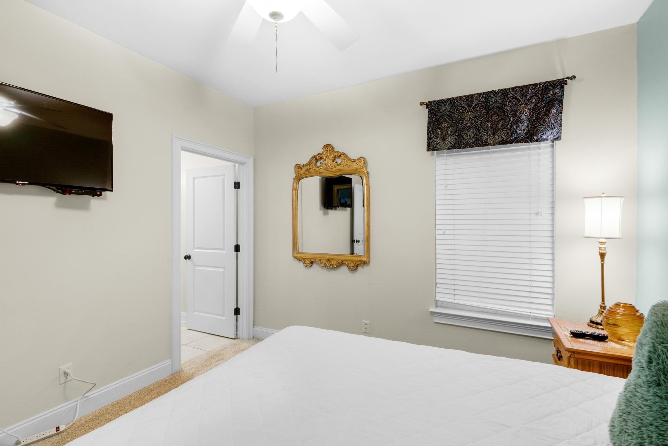 A bedroom with a bed, a television mounted on the wall, a mirror, a window with blinds, and a bedside table with a lamp.