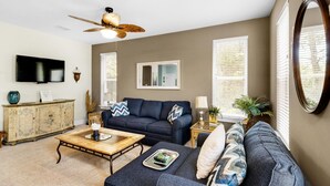 A living room with a dark blue sofa, a wooden coffee table, a ceiling fan, a television, and a decorative mirror. - Topsail Village 211- Sandy Feet Retreat (Santa Rosa Beach)