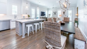 A modern kitchen with white cabinetry, a central island with stools, and a dining area with a wooden table and wicker chairs. - The Graytest 30A Beach House (Santa Rosa Beach)