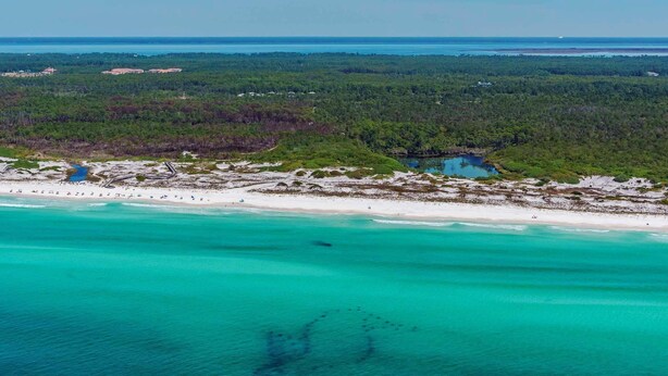 Vue aérienne d’une zone côtière avec une plage, des eaux turquoise et limpides, et une étendue de forêt dense.