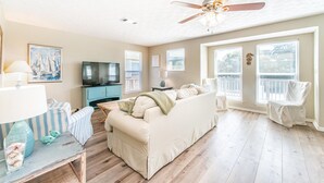 A spacious living room with a beige sofa, a television, a ceiling fan, and large windows. - Surfer Girl (Santa Rosa Beach)