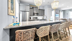 A modern kitchen with a granite countertop, stainless steel appliances, wicker bar stools, and a sign that reads "OYSTERS". - Sandy Cheeks (Santa Rosa Beach)