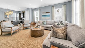 A spacious living room with a grey sofa, a wooden armchair, and a round coffee table. A kitchen is visible in the background. - Sandy Cheeks (Santa Rosa Beach)
