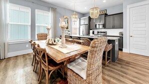 A dining area with a wooden table and wicker chairs, a kitchen with dark cabinets, and a white door. - Sandy Cheeks (Santa Rosa Beach)