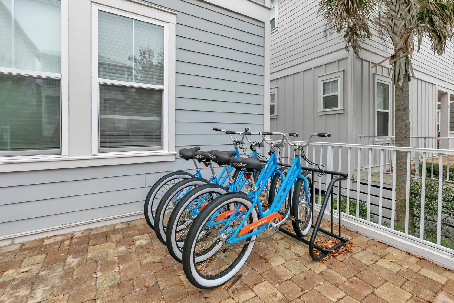 A row of blue bicycles parked against a building with white windows and a brick patio.