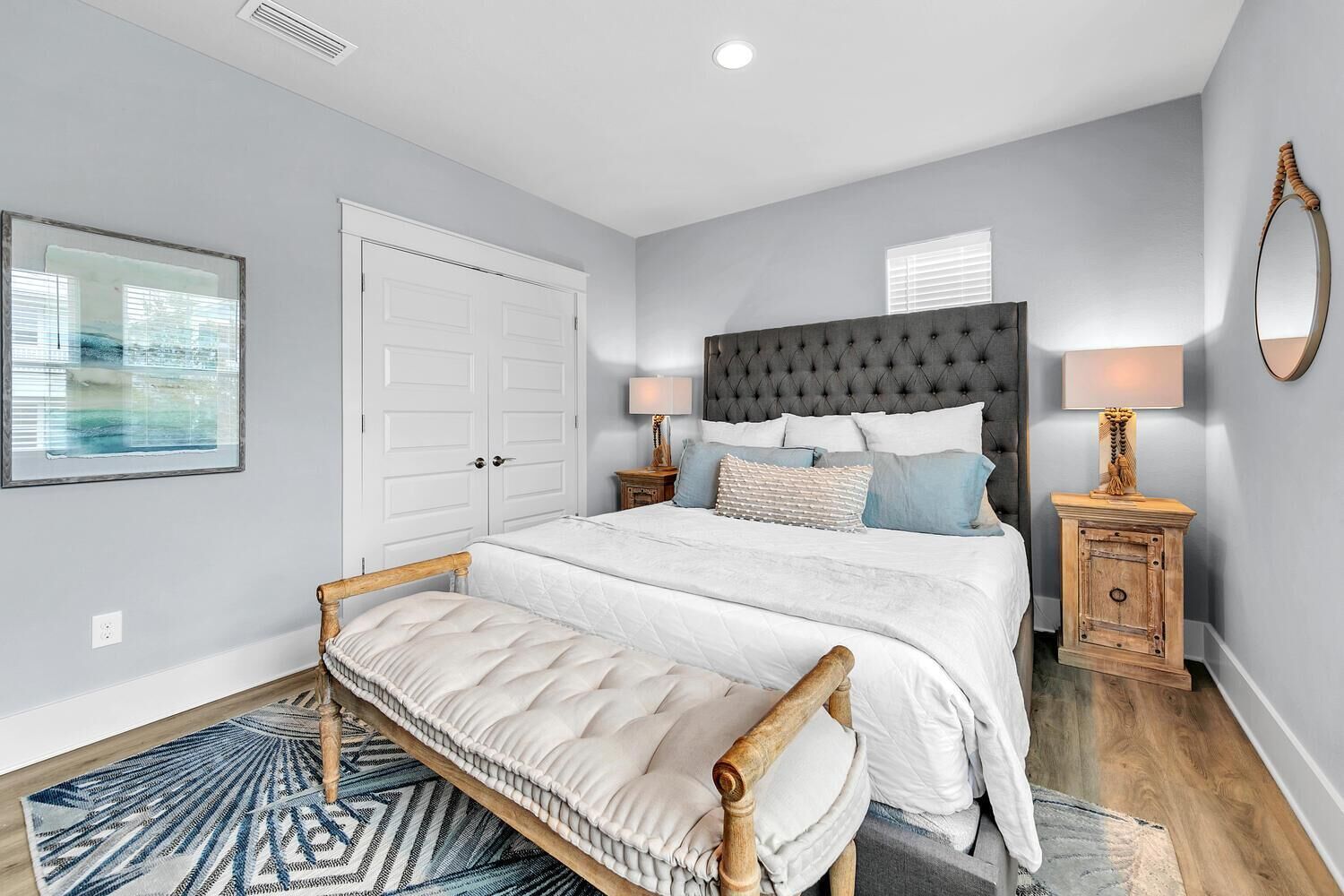 A bedroom with a large bed, a bedside table, a nightstand, a headboard, a door, a window, and a rug.