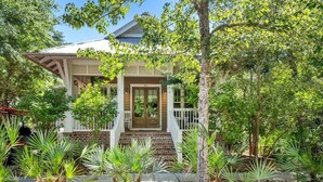 Une maison à deux étages avec un porche à l’avant, entourée de différentes plantes et d’arbres.