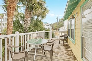 A wooden deck with a glass table and chairs, surrounded by a white railing, palm trees, and a building with a green awning. - Heron's Watch (Santa Rosa Beach)
