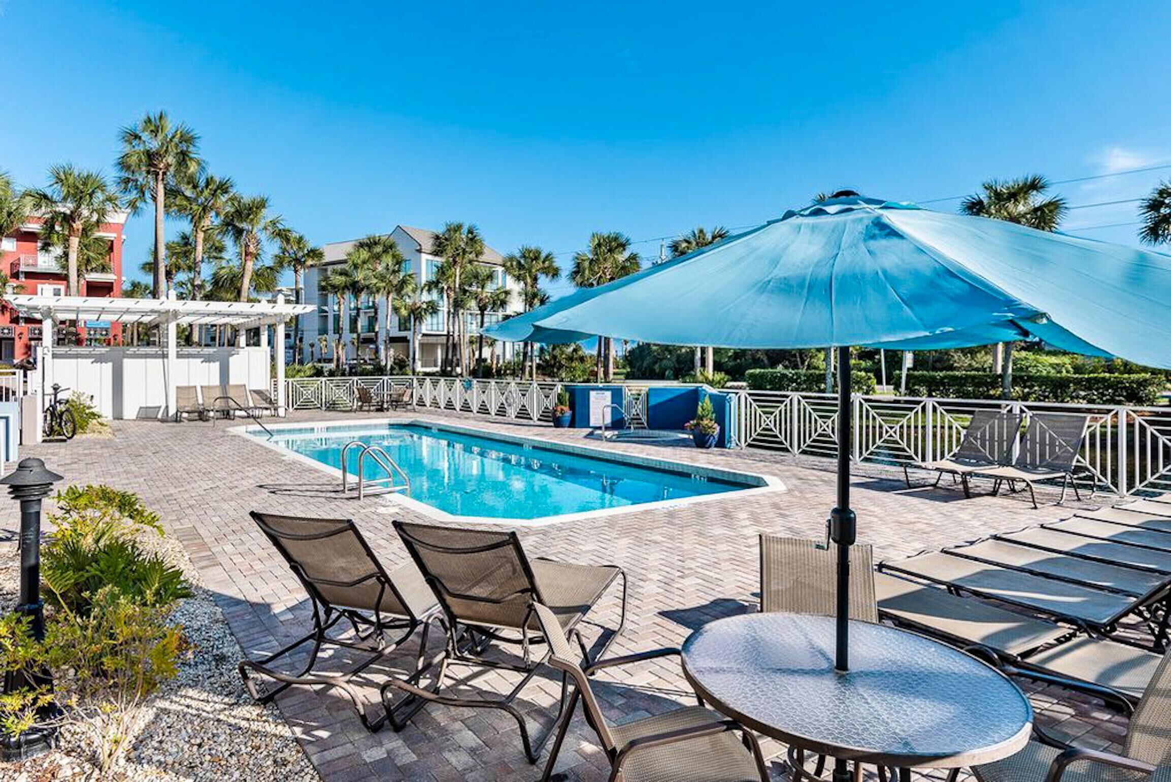 A pool area with lounge chairs and umbrellas, surrounded by palm trees and buildings.
