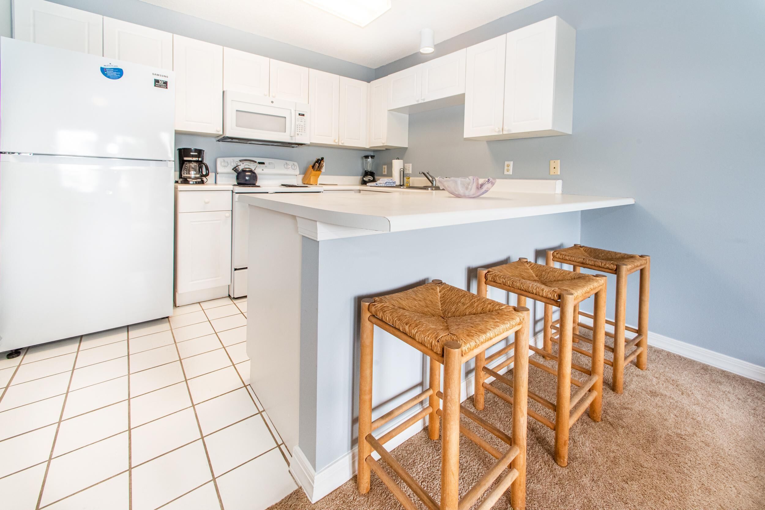 A modern kitchen with white cabinets, a white refrigerator, a microwave, and a coffee maker. There are three wooden stools with woven seats.