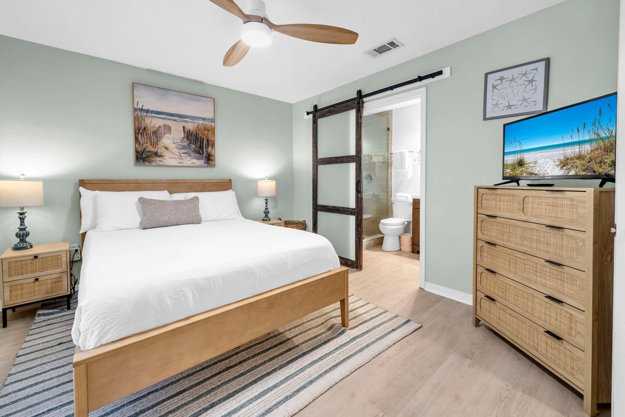 Un dormitorio con cama de madera, mesitas de noche, ventilador de techo, televisor y puerta corredera de vidrio que da al baño.