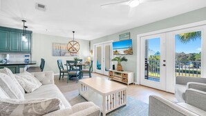 A spacious living room with a sofa, a coffee table, and a dining area with chairs and a table. There is a kitchen area with cabinets and a countertop. The room has large sliding glass doors leading to a balcony with a view of the outdoors.