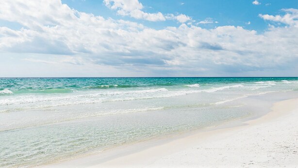 A beach with clear blue water and white sand under a partly cloudy sky.