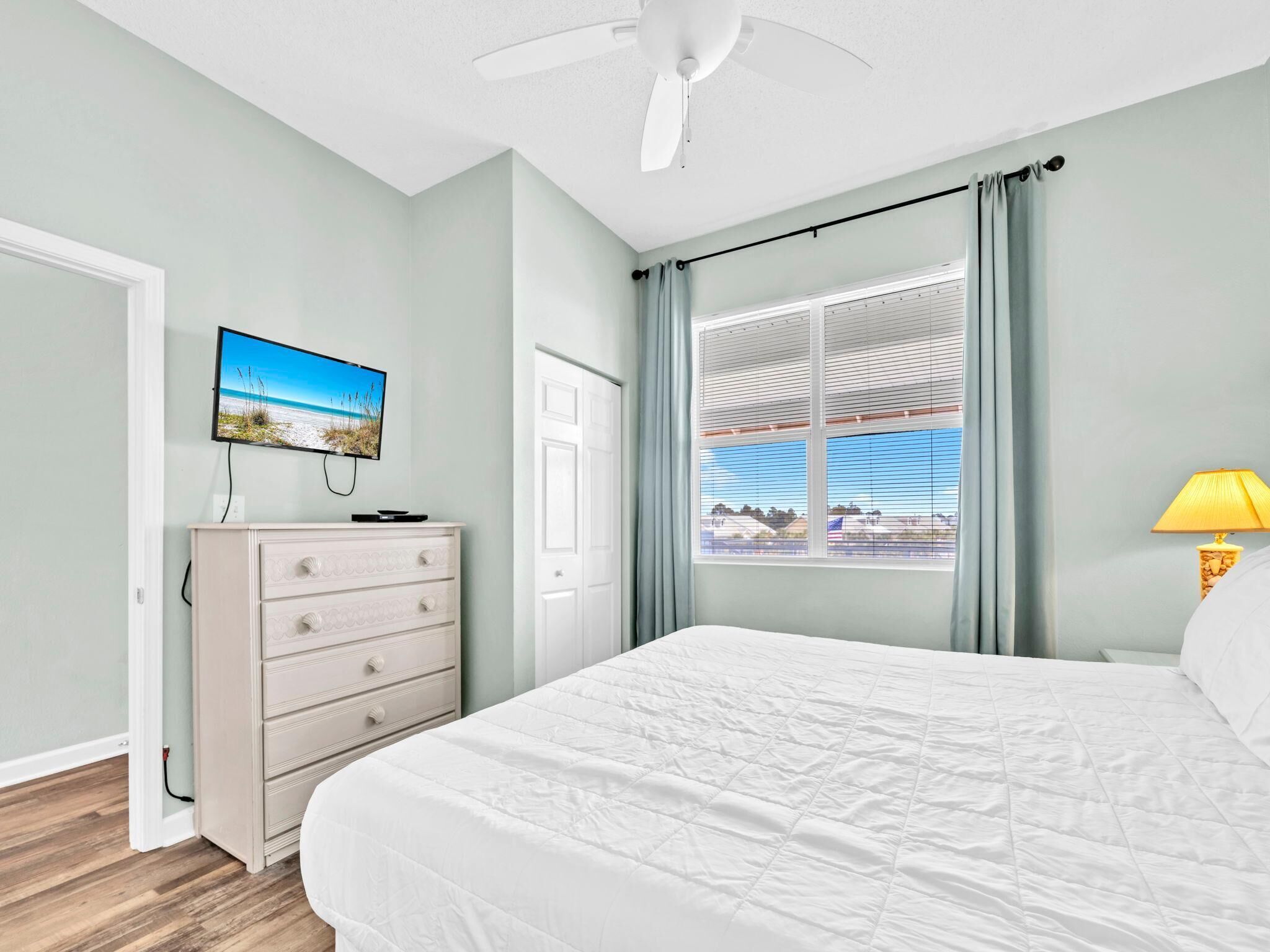 A bedroom with a bed, a dresser, a television on the wall, and a window with curtains.