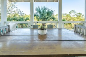 A covered outdoor dining area with a wooden table and chairs, a potted plant in the center, and a view of trees and foliage. - Day Is Done (Santa Rosa Beach)