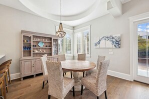 A dining area with a round wooden table, woven chairs, a wooden cabinet with glassware, and a large window with blinds. - Coastal Joy on 30A (Santa Rosa Beach)
