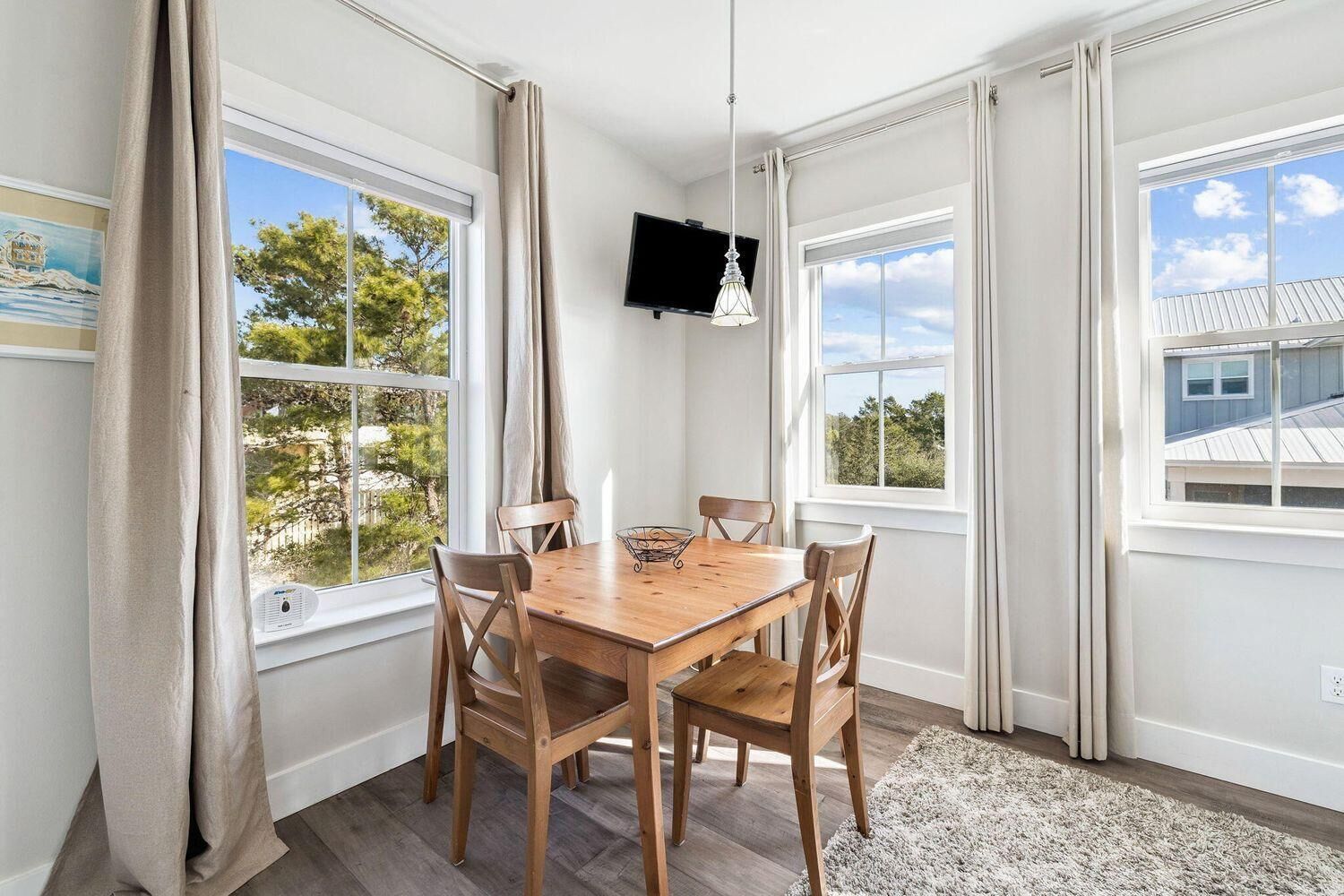 En spisestue med et træbord og stole, store vinduer med gardiner, et fladskærms-tv monteret på væggen og en loftlampe.