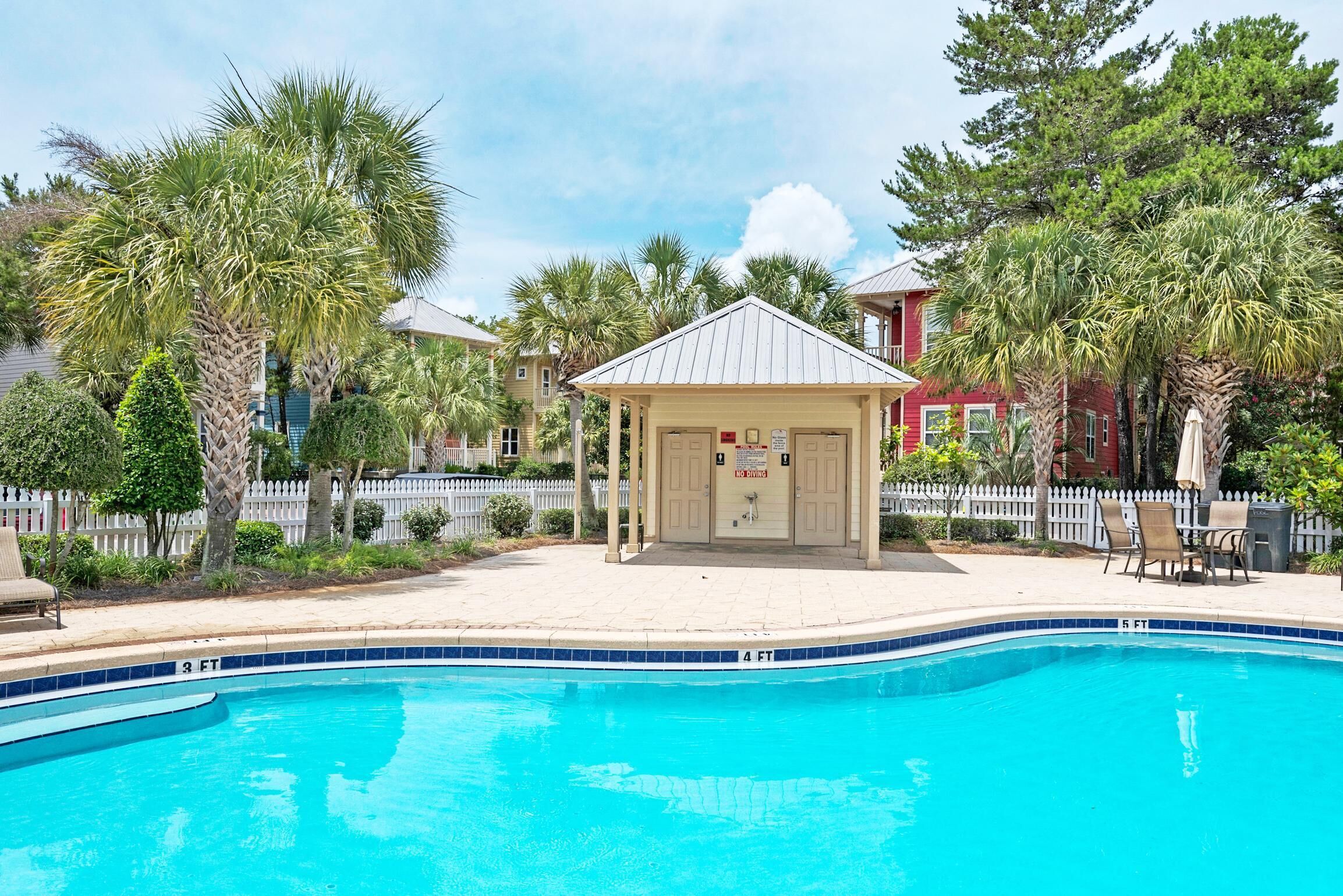 A swimming pool with a covered area, surrounded by palm trees and a white picket fence.