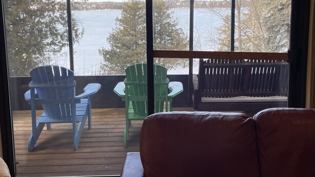 A view from a window overlooking a body of water, with a wooden deck, two blue Adirondack chairs, and a green Adirondack chair. - Relax and Rejuvinate at the cottage.  Get some forest therapy. (Bridgenorth)