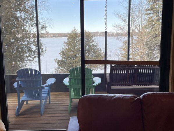 Une vue depuis une fenêtre donnant sur une étendue d’eau, avec une terrasse en bois, deux chaises Adirondack bleues et une chaise Adirondack verte.