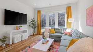Ein modernes Wohnzimmer mit einem weißen Sofa, einem weißen Couchtisch und einem großen Flachbildfernseher an der Wand. Zwei Fenster mit gelben Vorhängen, ein Deckenventilator und eine Pflanze auf dem Boden sind ebenfalls vorhanden.