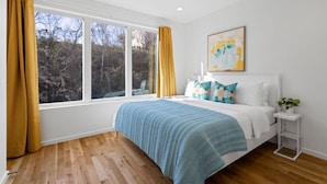 Ein Schlafzimmer mit großem Fenster, einem Bett mit blauer Bettdecke und einem weißen Nachttisch mit Pflanze.