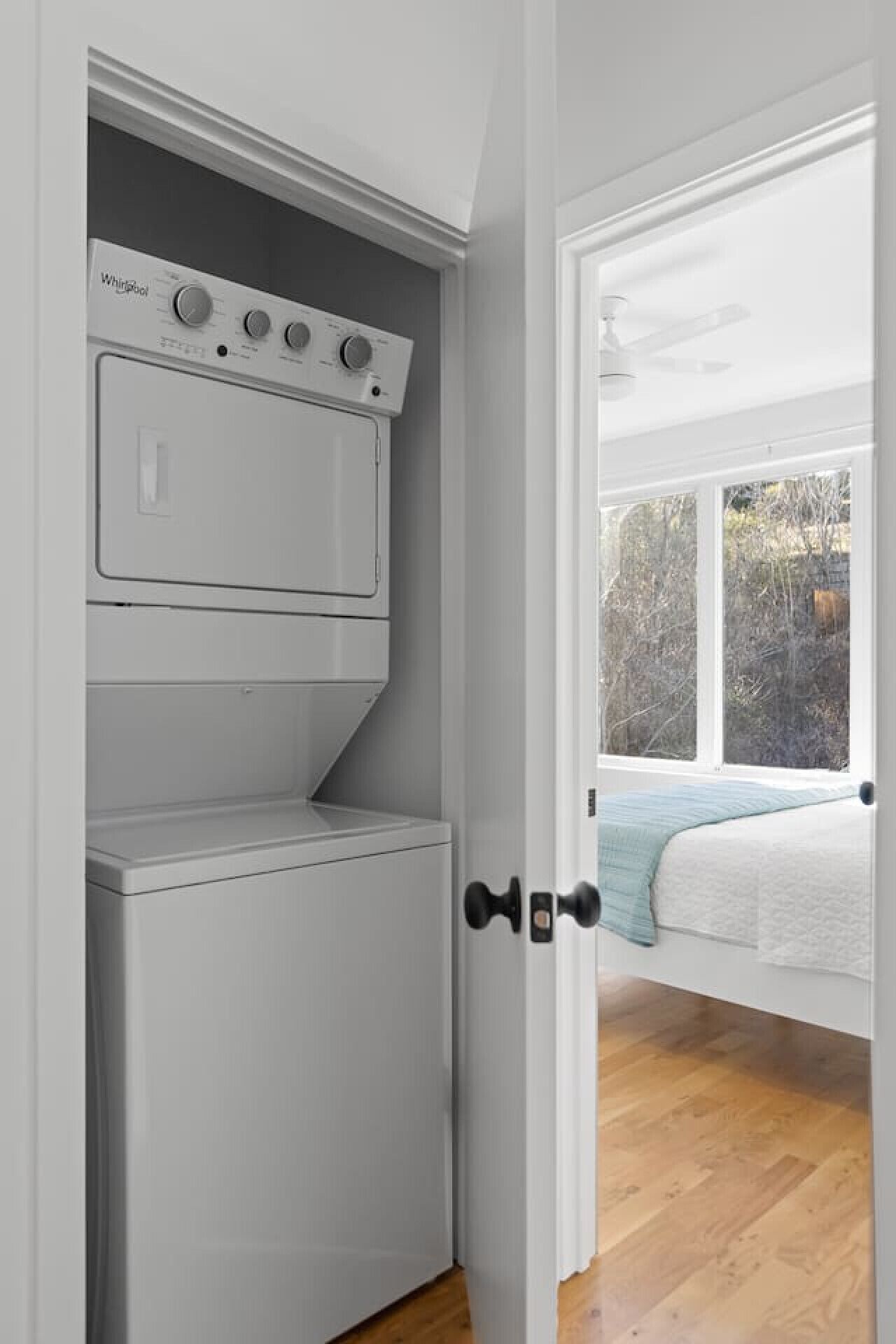 A laundry room with a front-loading washer and dryer, a door leading to a bedroom with a bed and a view of trees.