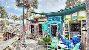 A wooden deck with green Adirondack chairs, a small table, and a tiki bar with colorful decorations. - Chalet Mer (Santa Rosa Beach)