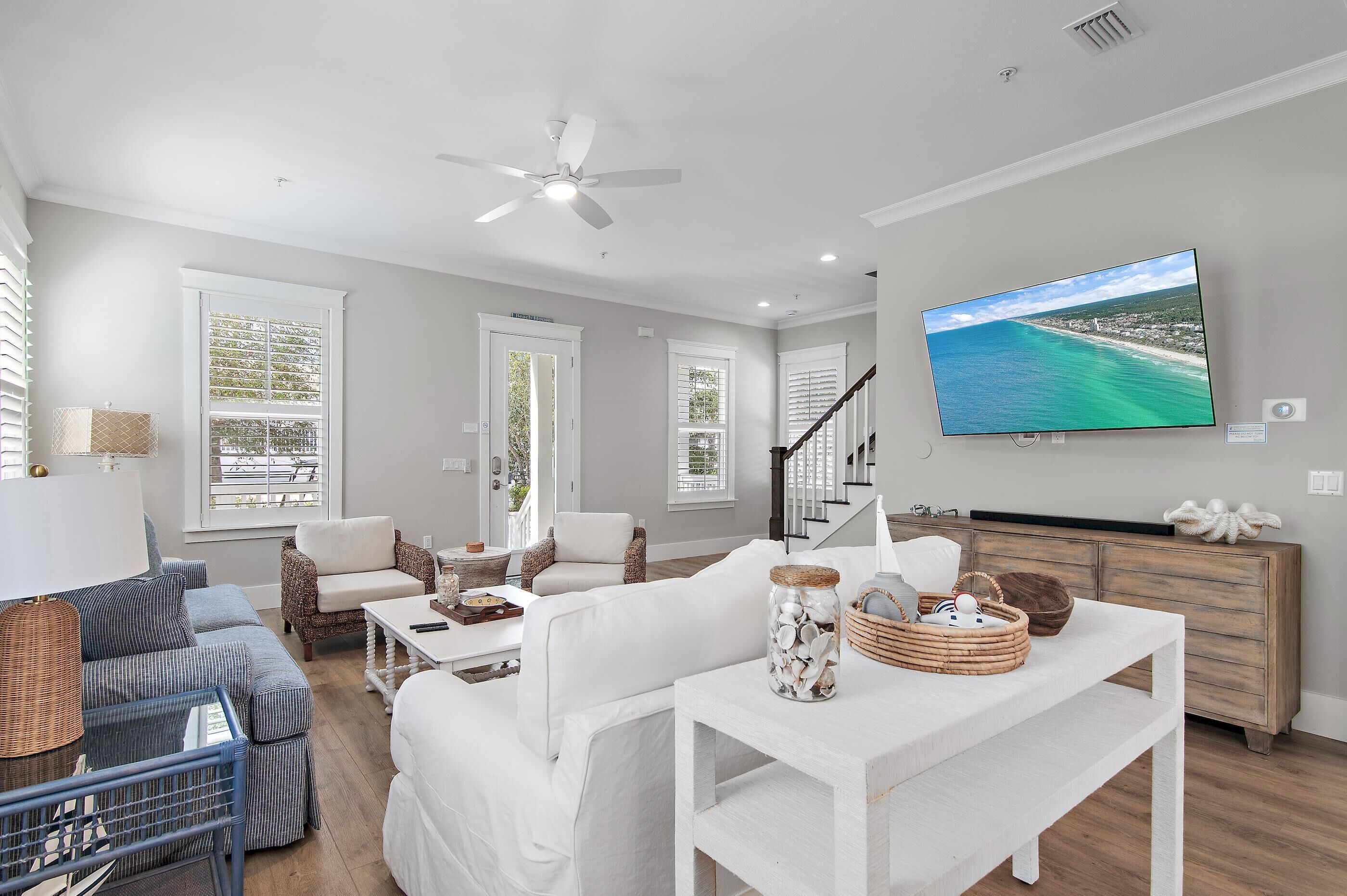 A spacious living room with a large flat-screen TV, white furniture, and a view of the ocean through the windows.