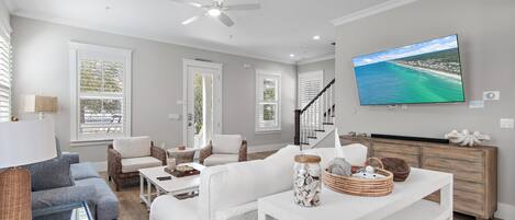 A spacious living room with a large flat-screen TV, white furniture, and a view of the ocean through the windows.