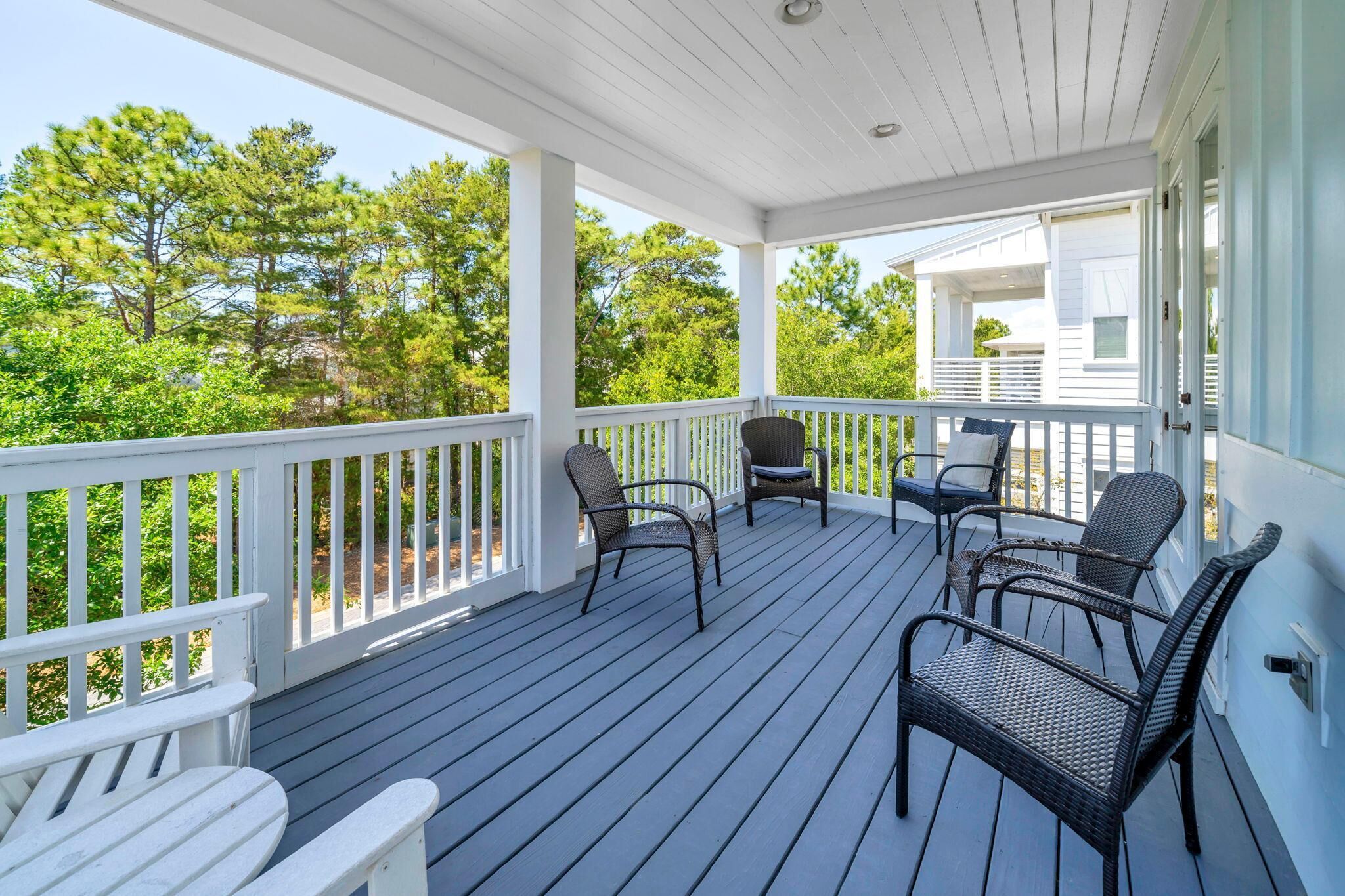 A covered porch with white railings, wicker chairs, and a view of trees and a house.