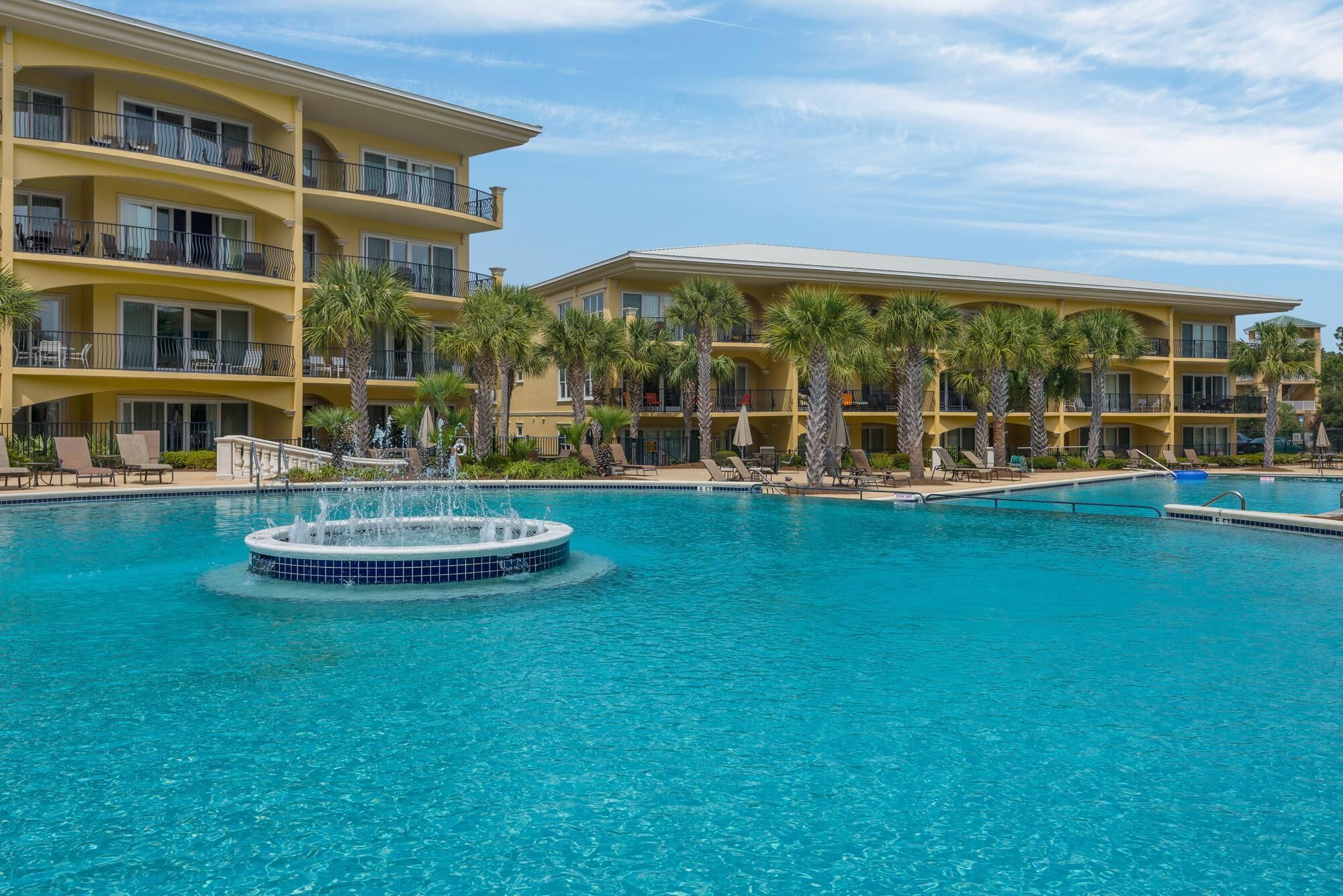 A hotel with multiple balconies, a central pool with a fountain, and palm trees.