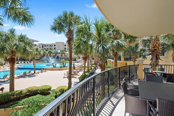 A poolside area with lounge chairs and palm trees, a dining area with tables and chairs, and a building with arched windows in the background.