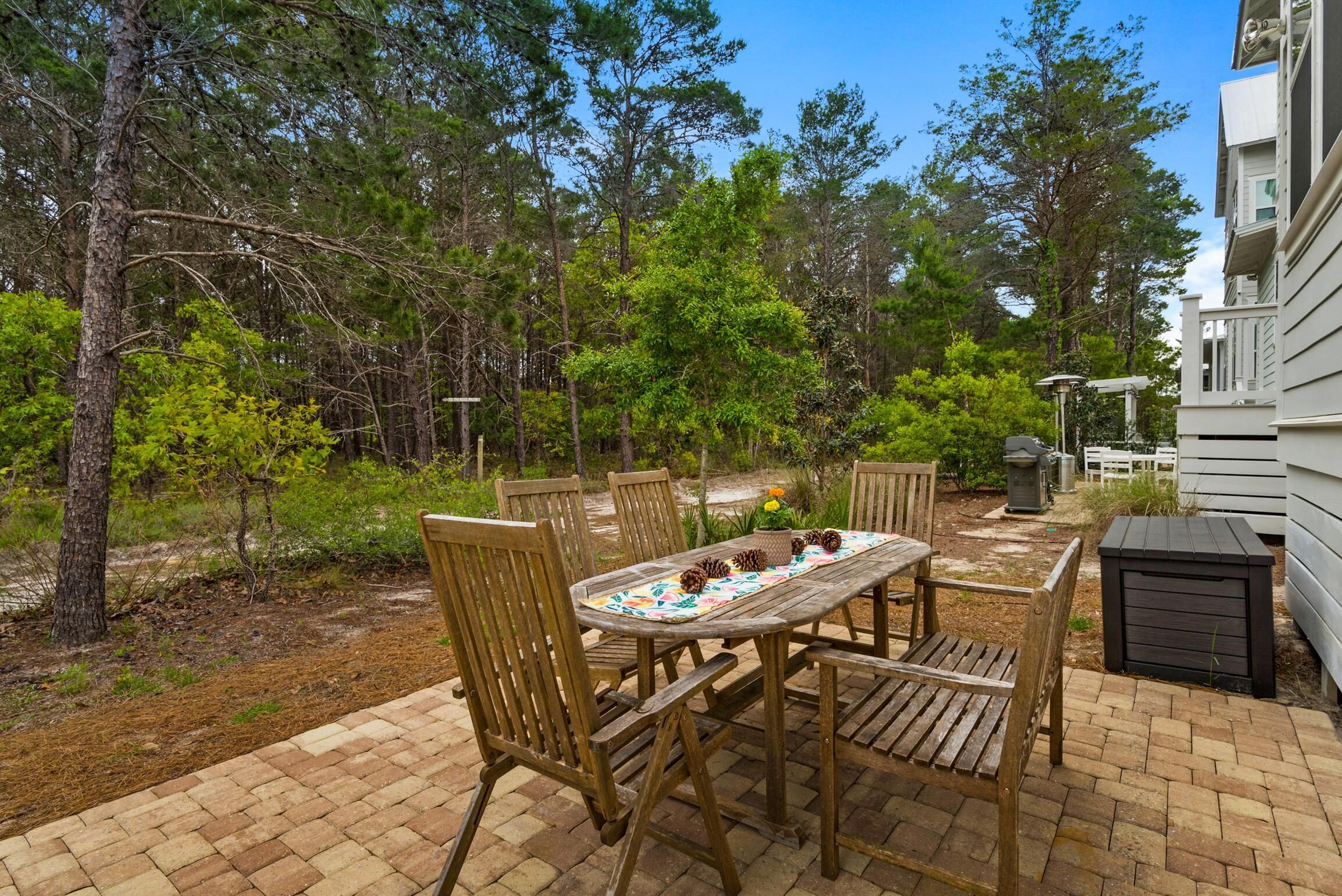 An outdoor patio with a wooden table and chairs, surrounded by trees and a brick pathway.