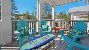 A covered porch with turquoise Adirondack chairs, a surfboard with text, and a view of trees and houses. - 30A Sunshine Shack (Santa Rosa Beach)