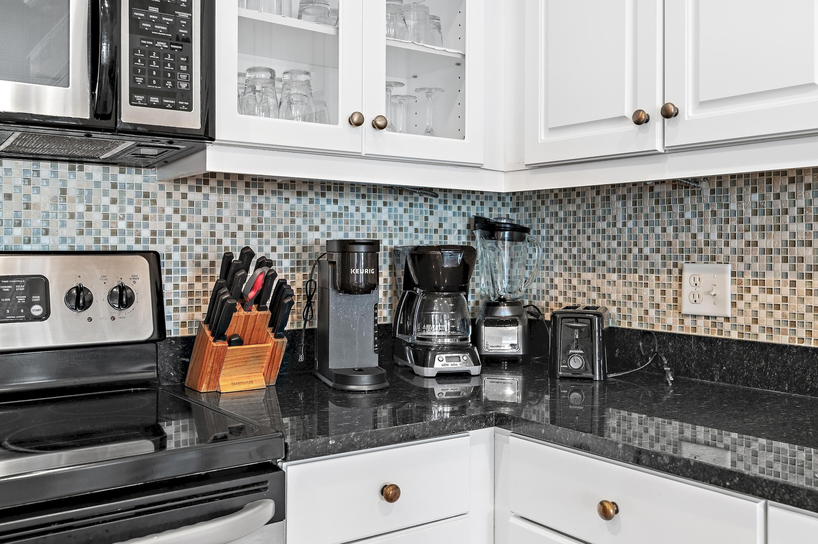 Una cocina con armarios blancos, encimera negra, estufa de acero inoxidable, cafetera y licuadora.
