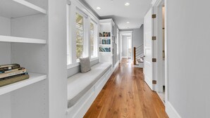 A hallway with white walls, wooden flooring, built-in bookshelves, a bench with a cushion, and a staircase. - The Atticus (Panama City Beach)