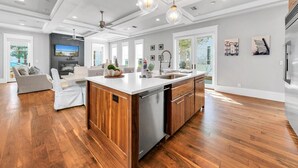 A spacious kitchen with a central island, wooden cabinets, and a dishwasher. There is a dining area with a sofa and a TV. The room has large windows and a ceiling fan. - The Atticus (Panama City Beach)
